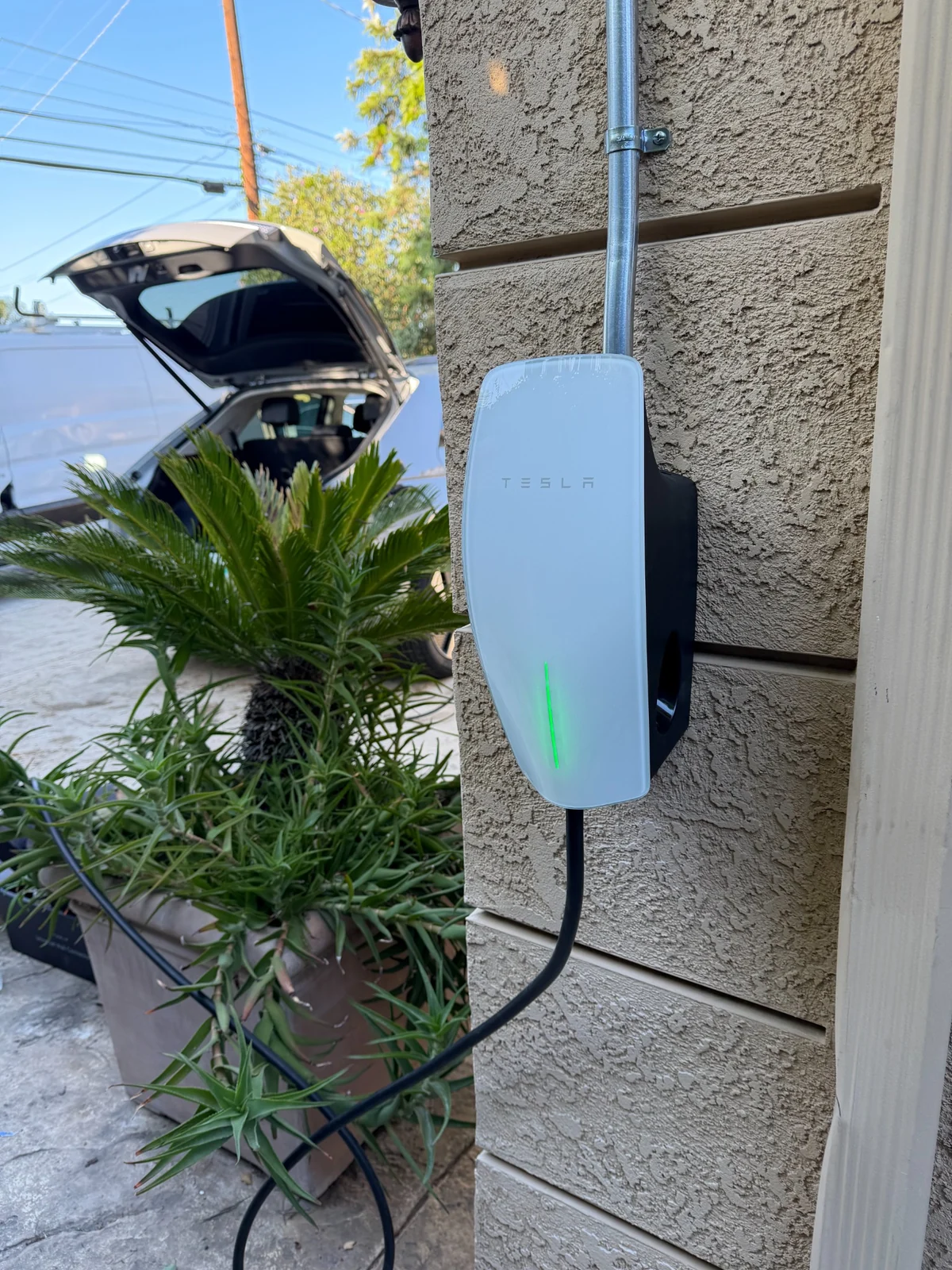 Tesla Wall Connector mounted outdoors on a stucco wall beside a driveway.